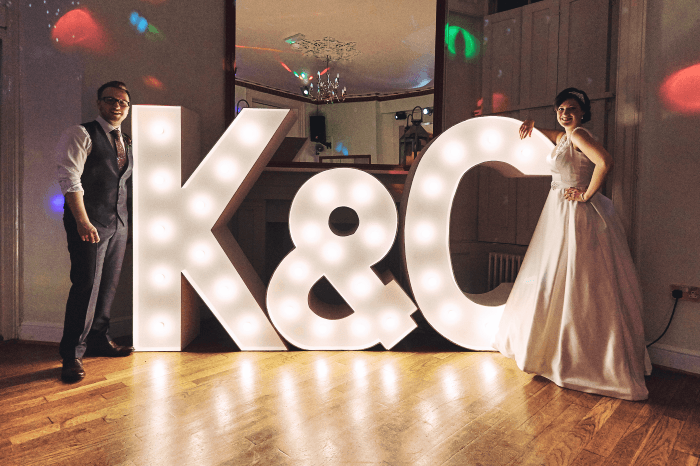 Идея свадебной съемки: Пара стоит у гигантских светящихся инициалов на приеме