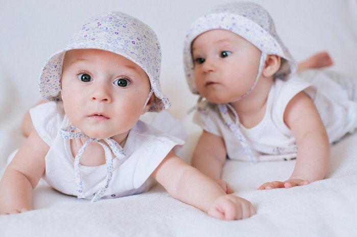 фотосессия новорожденных близнецов: новорожденные близнецы в одинаковых нарядах и шапочках лежат на животе