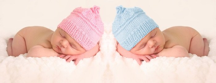 фотографии новорожденных близнецов: новорожденные близнецы в розовых и голубых вязаных шапочках позируют, положив головы на скрещенные руки