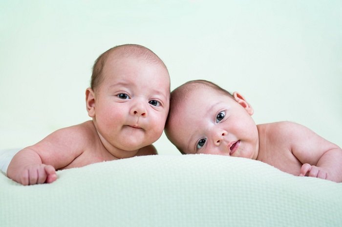 фотография новорожденных близнецов: новорожденные близнецы на животе смотрят на камеру, нежно положив головы вместе