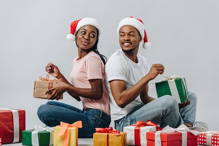 Идеи для рождественской фотосессии: пара в шапках Санты сидит спина к спине и вместе открывает подарки