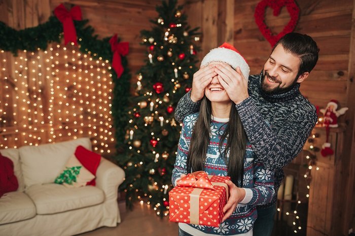 Советы по фотосессии рождественской пары: мужчина закрывает глаза подруги руками, чтобы она не видела подарок, который держит в руках
