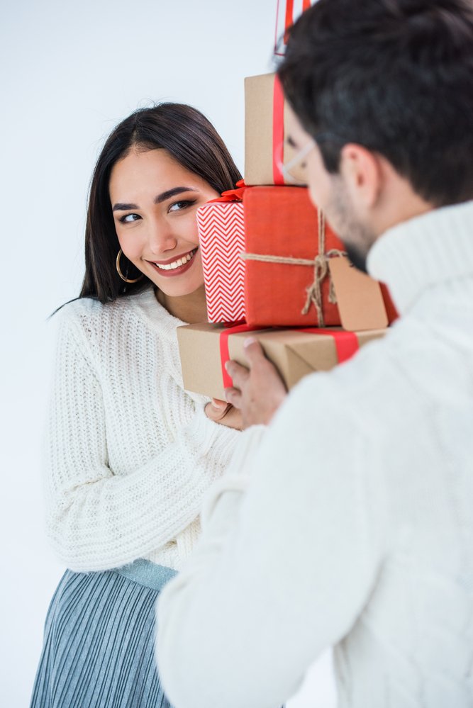 Советы по фотосессии рождественской пары: женщина смотрит на своего парня из-за упакованных рождественских подарков
