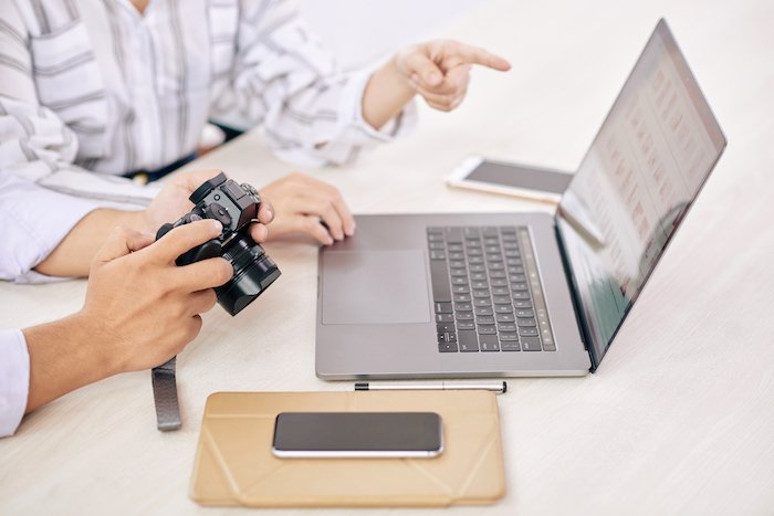 Снимок фотографа с камерой и смартфоном, использующего ноутбук с программой для управления фотографиями