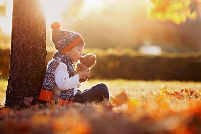 Ребенок с плюшевым мишкой, сидящий у ствола дерева, как пример детской фотографии