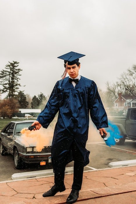 Уникальная идея выпускной фотографии выпускника школы в шапочке и мантии, держащего в руках цветные дымовые шашки